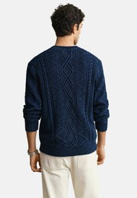 Man draagt een marineblauwe kabelgebreide trui en crèmekleurige broek, gezien van achteren tegen een effen achtergrond.