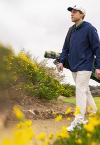 Niebieska, lekka kurtka z kapturem, białe spodnie, trzymający w ręku koc w kratę, noszący kije golfowe, spacerujący po trawiastej ścieżce z żółtymi kwiatami.