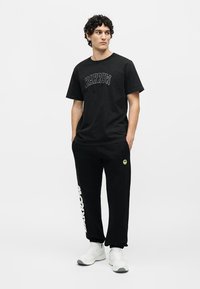Homme portant un t-shirt noir "BARROW", un pantalon de jogging noir avec logo et texte, des baskets blanches, debout les mains dans les poches, regardant sur le côté.