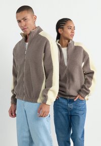 Fleece jacks met een rits aan de voorkant, met een bruin lichaam en crèmekleurige mouwen. De textuur is zacht en weelderig, geschikt voor casual kleding.