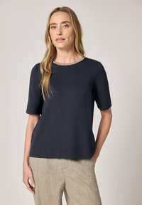 Kobieta o długich blond włosach, ubrana w granatową bluzkę z krótkim rękawem z kontrastowym dekoltem oraz beżowe spodnie o fakturowanej powierzchni, stojąca na jednolitym tle.