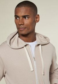 Light beige zip-up hoodie with a drawstring hood, featuring a soft texture and white zipper details. Worn over a white t-shirt.