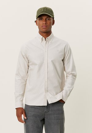 Chemise à rayures en tissu beige clair et blanc cassé, avec des manches longues et un col légèrement structuré. Associée à une casquette verte.