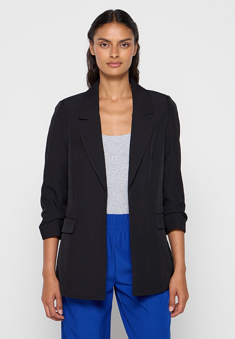 Blazer negro con un diseño a medida, solapas muesca y mangas remangadas. Lleva puesto sobre una blusa gris, combinado con pantalones azules.