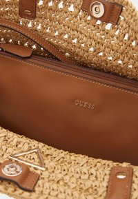 Bolso de paja tejido con un interior de cuero marrón, que cuenta con un bolsillo con cremallera y un logotipo en tono dorado. Destacado con un detalle triangular.