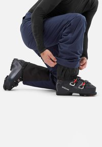 Bottes de ski noires avec ferrures rouges, portées par une personne à genoux en pantalon bleu marine. Le pantalon a un ourlet resserré sur la botte, mettant en valeur un design élégant.