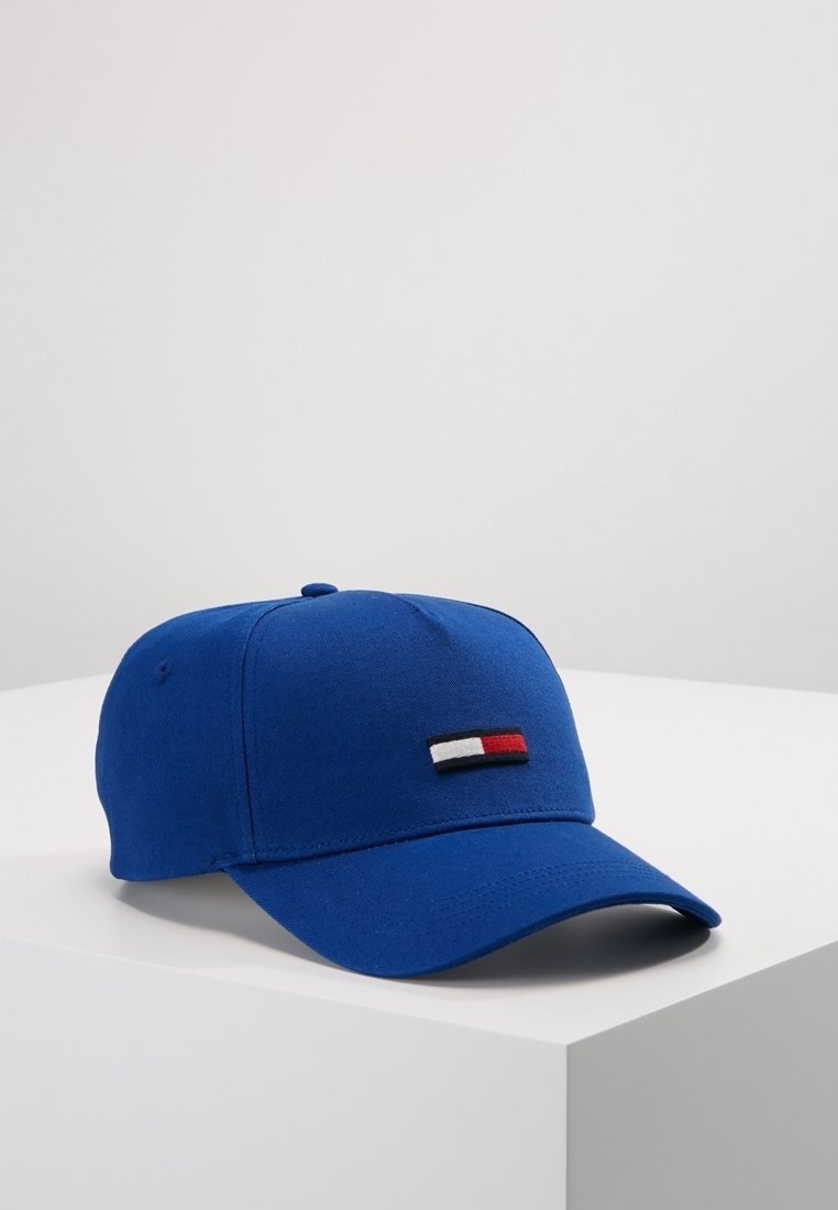 Casquette de baseball bleue avec une visière incurvée et un petit logo rectangulaire rouge, blanc et noir à l'avant, placée sur une surface blanche.