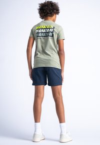 Lichtgroene katoenen T-shirt met bold zwart en gele "HAWAII" tekst op de rug, gecombineerd met navy shorts en witte sneakers.