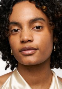 Gros plan sur une personne ayant des cheveux bouclés et foncés, avec un teint clair, portant de petites boucles d'oreilles en or. Elle a un teint lisse et un maquillage naturel.