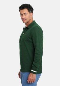 Camisa polo de manga larga en verde oscuro con cuello abotonado, adornada con franjas de acento blancas en los puños y un sutil logo en el pecho.