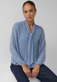 Hellblaue Bluse mit vertikalen Plisseefalten, einem gebundenen Ausschnitt und langen Ballonärmeln. Kombiniert mit dunklen Jeans, mit einer glatten, fließenden Textur.