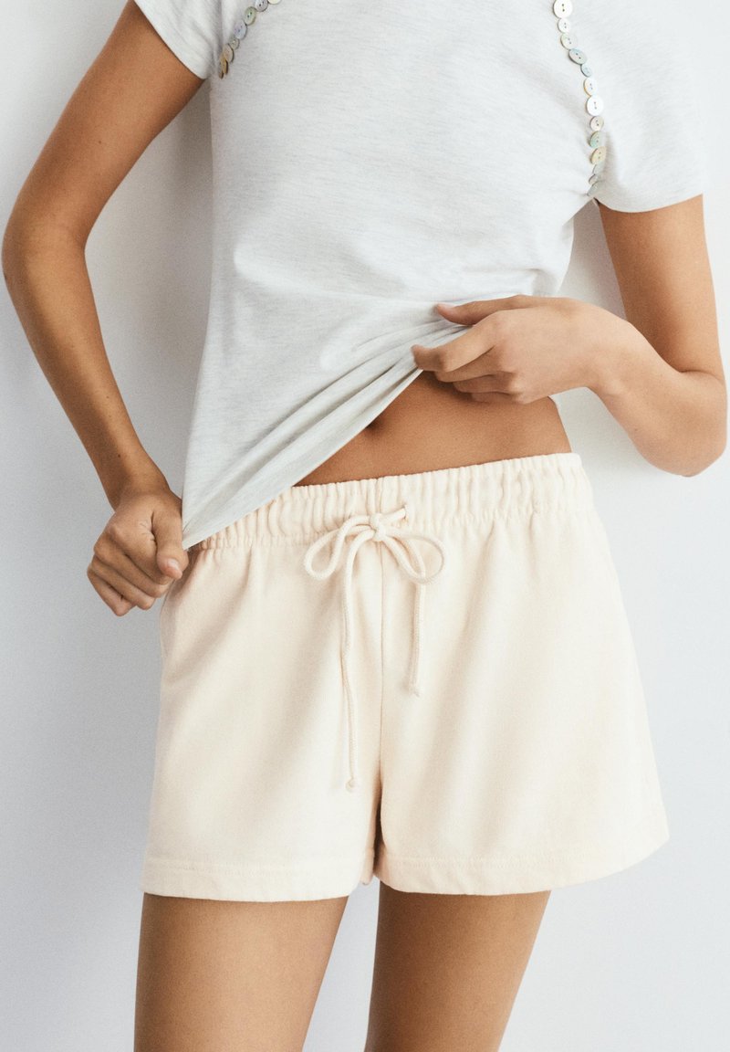Personne portant un short beige clair avec cordon et une chemise blanche à manches courtes, soulevant légèrement la chemise pour dévoiler la taille.