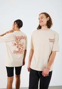 Beige oversized tričko s nápisom "Beautifully Broken" a grafickými motívmi anjela v hrdzavej farbe na chrbte a na ľavej hrudi; vyrobené z bavlny.
