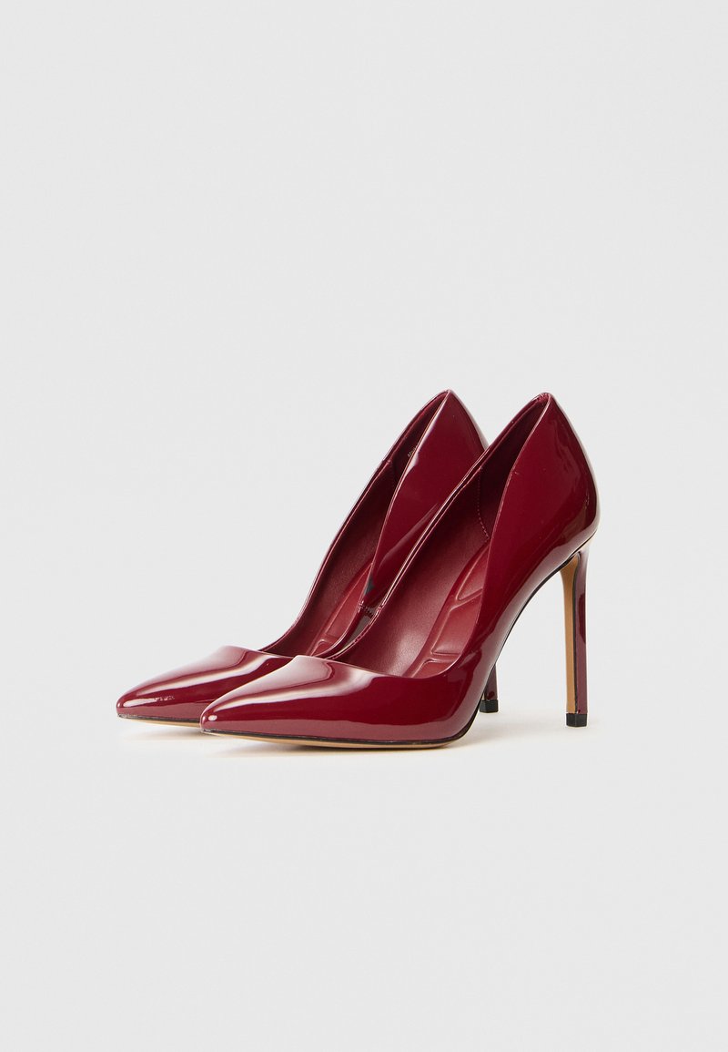 Décolleté in vernice bordeaux con punta affusolata, texture liscia e tacco a stiletto sottile. Dettagli interiori sottili visibili.