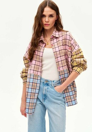 Kobieta o długich brązowych włosach nosi oversize'ową koszulę w kratę w kolorach różu, żółci i niebieskiego, biały top na ramiączkach oraz jasnoniebieskie dżinsy, z ręką w kieszeni.