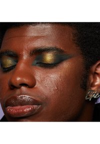 Gros plan d'un visage à la peau foncée, avec un maquillage des yeux noir et or audacieux, des lèvres brillantes et une boucle d'oreille "Epic". Peau texturée et sourcils bien définis.