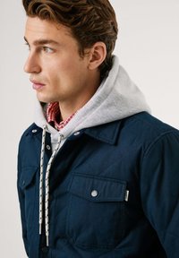 Chaquetón de navy acolchado con dos bolsillos en el pecho, sudadera con capucha gris y camiseta a cuadros debajo. Cuenta con un cierre de botones y cordones de contraste.
