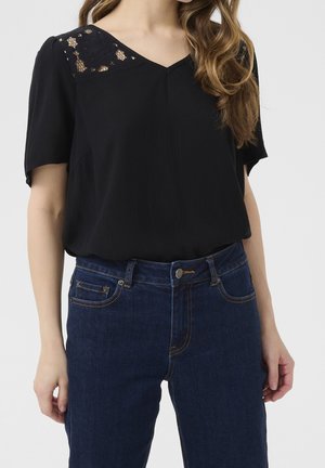 Femme portant un chemisier noir avec un détail en dentelle sur l'épaule et un jean bleu en denim, debout devant un fond gris clair.