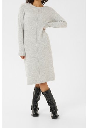 Femme portant une longue robe en maille gris clair à manches longues et des bottes en cuir noires mi-mollet, debout, une main sur la hanche.