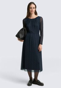 Marineblaues Kleid mit langen, transparenten Ärmeln, strukturiertem Muster, plissiertem Ausschnitt und passender schwarzer Tasche. Kombiniert mit schwarzen Schuhen und Strumpfhosen.