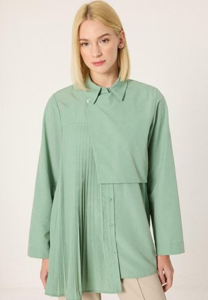 Ξανθιά γυναίκα που φοράει μια ασύμμετρη, σιφονένια πουκάμισο σε μέντα πράσινο χρώμα με κουμπιά και ένα φαρδύ καπάκι πάνω από το στήθος, στέκεται μπροστά σε ένα απλό φωτεινό φόντο.