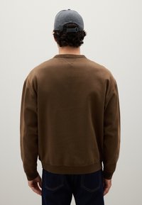 Felpa marrone con collo a giro e polsini a costine, indossata con jeans scuri. Un cappellino grigio è visibile, con un regolatore in plastica sul retro.