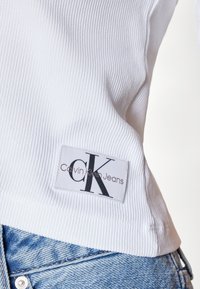 Valkoinen röyhelömäinen kangas, jossa on Calvin Klein Jeans -merkki mustalla tekstillä ja logolla. Vaate on stailattu vaaleansinisellä denimpäällä.