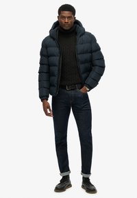 Donkerblauwe gewatteerde jas met een capuchon, met een gekreukt textuur, gecombineerd met een zwarte kabelgebreide trui, blauwe jeans en zwarte veterschoenen.