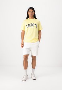 Gul bomull T-skjorte med marineblå "LACOSTE" trykk, kombinert med hvite shorts og hvite sneakers. Modellen står foran en nøytral bakgrunn.
