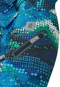 Blue and green patterned fabric with geometric dots, featuring a gray logo patch and a zippered pocket with teal accents.