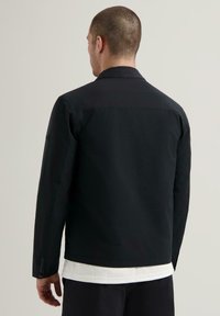 Giacca nera con un design colletto, tessuto texturizzato e dettagli con bottoni sulle maniche, abbinata a una camicia bianca sotto.