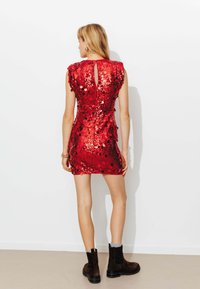 Robe rouge à sequins avec manches courtes, coupe ajustée, détail dos en trou de serrure, associée à des bottines marron et des accents de chaussettes grises.