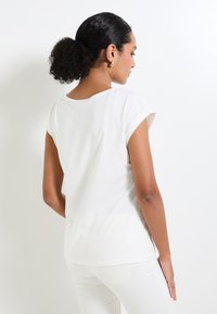 T-shirt ajusté à manches courtes blanc en tissu doux, avec un col rond et un dos légèrement creusé. Couture visible.