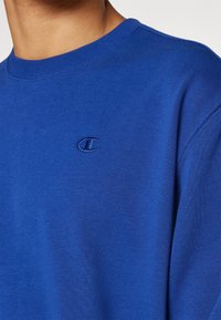 Blå bomullströja med rundad halsringning. Har en broderad logotyp på vänster bröst och en mjuk textur.