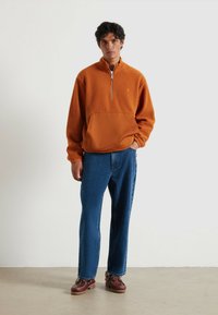 Pullover arancione in pile con zip a metà, superficie texturizzata e pannello inferiore uniforme, abbinato a jeans blu a gamba dritta e scarpe marroni.