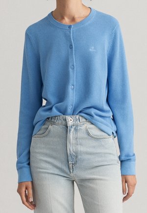 Femme portant un cardigan bleu clair texturé boutonné avec un jean taille haute délavé sur un fond neutre.