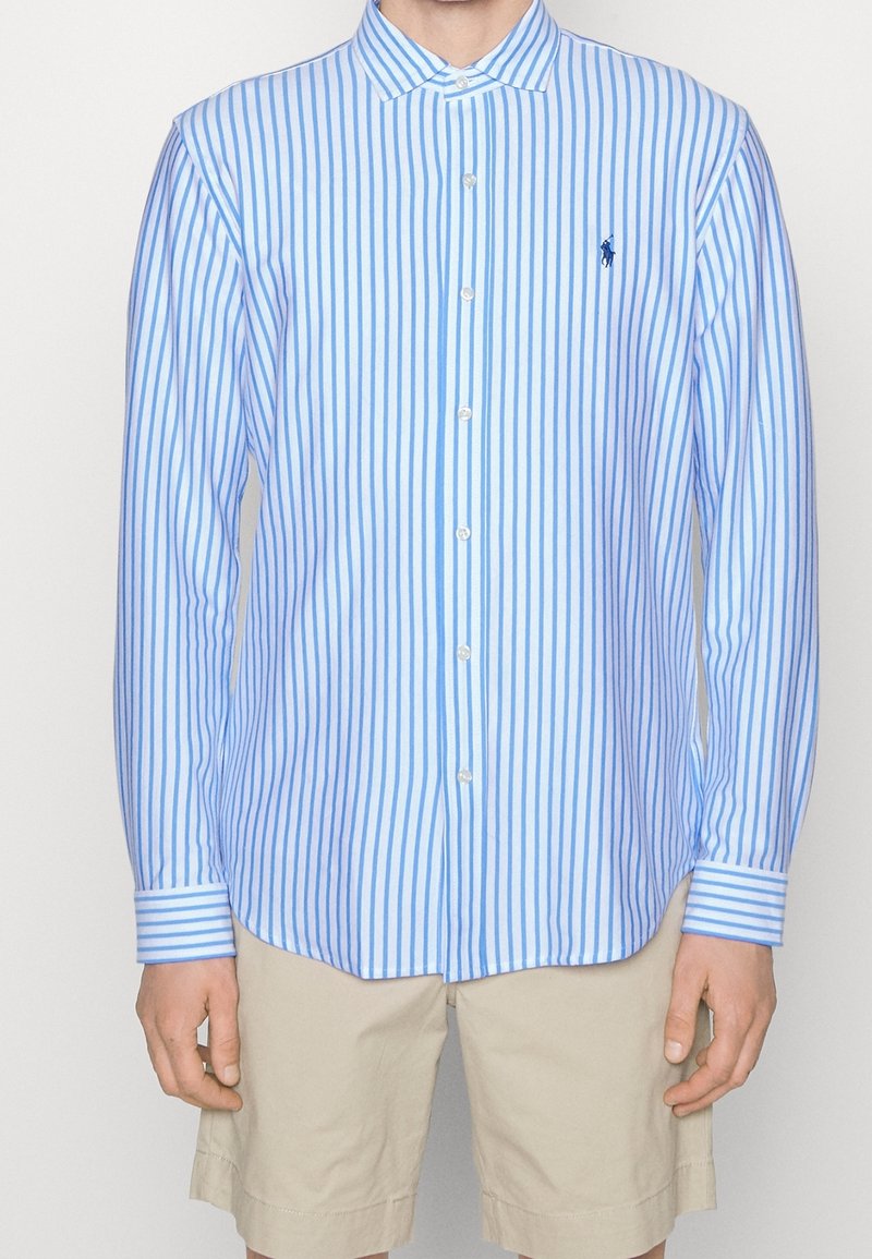 Homme portant une chemise à manches longues à rayures verticales bleues et blanches, boutonnée, avec un short beige, debout devant un fond uni.