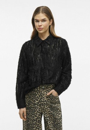 Zwarte blouses met textuur, voorzien van een kraag en lange mouwen. Gecombineerd met brede, beige broek met luipaardprint.