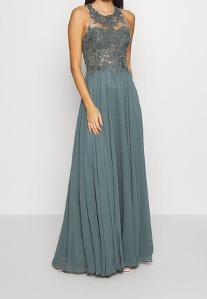 Hihaton lattiapitkä liiturinharmaa mekko, jossa on läpikuultava koristeltu pitsiyläosa ja laskeutuva laskostettu hame, pukeutuneena seisovalla henkilöllä.