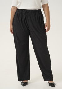 Schwarze plissierte Hose mit elastischem Bund, die locker sitzt und sich leicht zum Knöchel verjüngt, kombiniert mit spitzen schwarzen Schuhen.