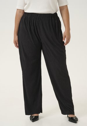Pantaloni neri plissettati con cintura elastica, caratterizzati da una vestibilità ampia che si stringe leggermente verso la caviglia, abbinati a scarpe nere a punta.