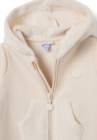 Chaqueta color crema con cremallera, capucha de peluche, bolsillo frontal y diseño bordado de oso. Hecha de un material suave y acogedor.