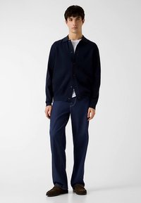 Cardigan blu navy con chiusura a bottoni, indossato sopra una T-shirt bianca. Abbinato a jeans in denim scuro. Scarpe marroni con texture completano l'outfit.