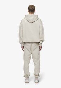 Beige hettegenser og matchende sweatpants laget av myk stoff, med en avslappet passform, ribbede mansjetter og hette. Hvite joggesko fullfører antrekket.