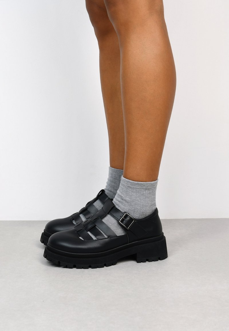 Sandales en cuir noir avec plusieurs brides, une semelle épaisse et un accent de boucle, portées avec des chaussettes grises.