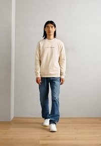 Šviesiai rudas džemperis su mažo teksto raštu, mėlyni džinsai ir balti sportbačiai. Aplinkoje matoma šviesiai pilka siena ir medinis grindys.