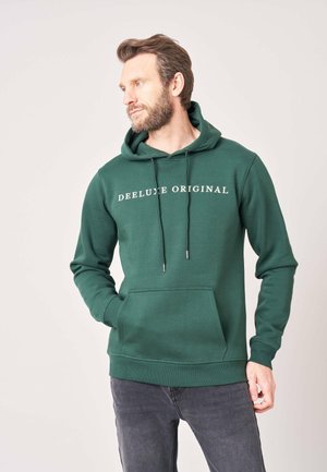 Man met baard die een groene hoodie draagt met de tekst "Deeluxe Original", handen in de zakken, kijkt opzij tegen een effen achtergrond.