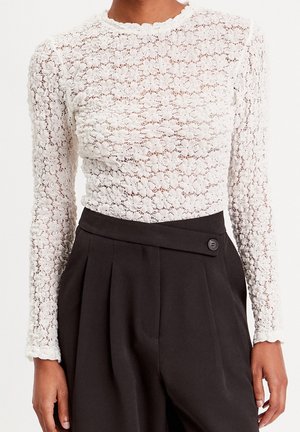 Femme portant un haut à manches longues en dentelle florale blanche associé à un pantalon noir plissé taille haute avec un détail de ceinture boutonnée.