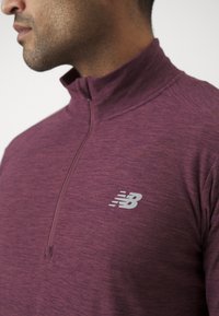 Bordo pulover s četvrtim zipom izrađen od teksturirane tkanine, s diskretnim sivim logotipom "New Balance" na lijevoj strani prsnog koša.