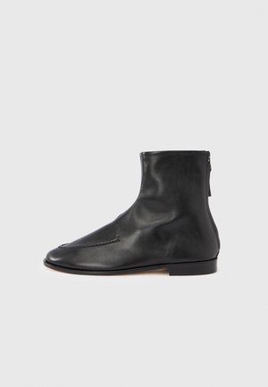 Stivaletto alla caviglia in pelle nera con finitura liscia, punta appuntita, dettagli di cuciture minime e piccola cerniera posteriore per la chiusura. Tacco piatto.